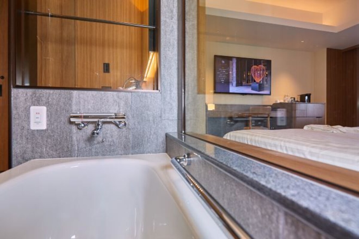 A modern bathroom with a large bathtub, a wooden vanity, and a mirror reflecting a bedroom with a bed and a TV.