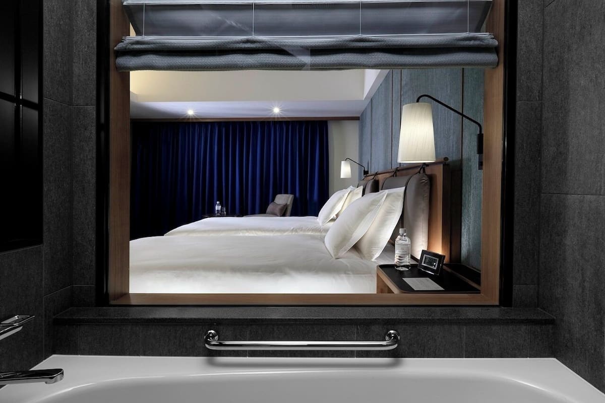 A modern hotel room with a large bed, a bedside table with a lamp, a mirror, and a view of a blue curtain.
