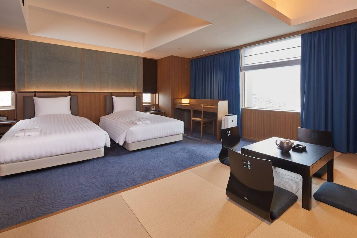 A hotel room with two beds, a desk, a chair, a table, and a view of the city.