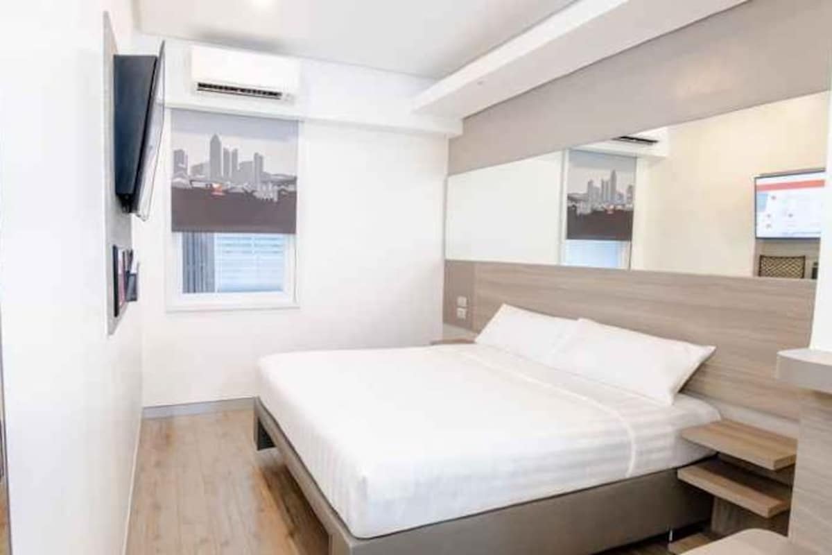 Habitación de hotel moderna con una cama, televisor montado y ventana con vista a la ciudad.