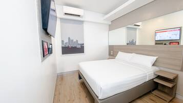 A modern hotel room with a bed, a TV, an air conditioning unit, a cityscape poster, and a digital screen.