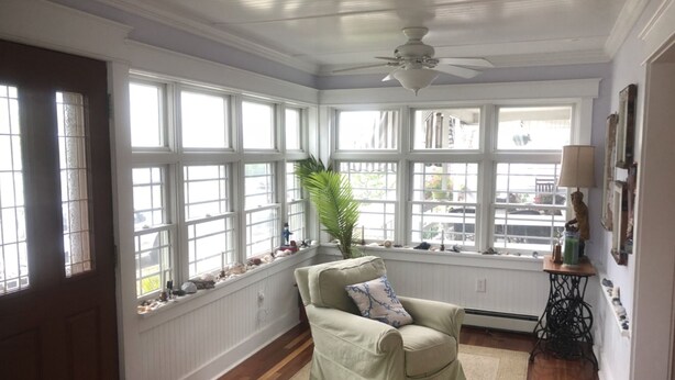 Una habitación con un amplio ventanal, un ventilador de techo, un sillón verde y una mesita con una lámpara.