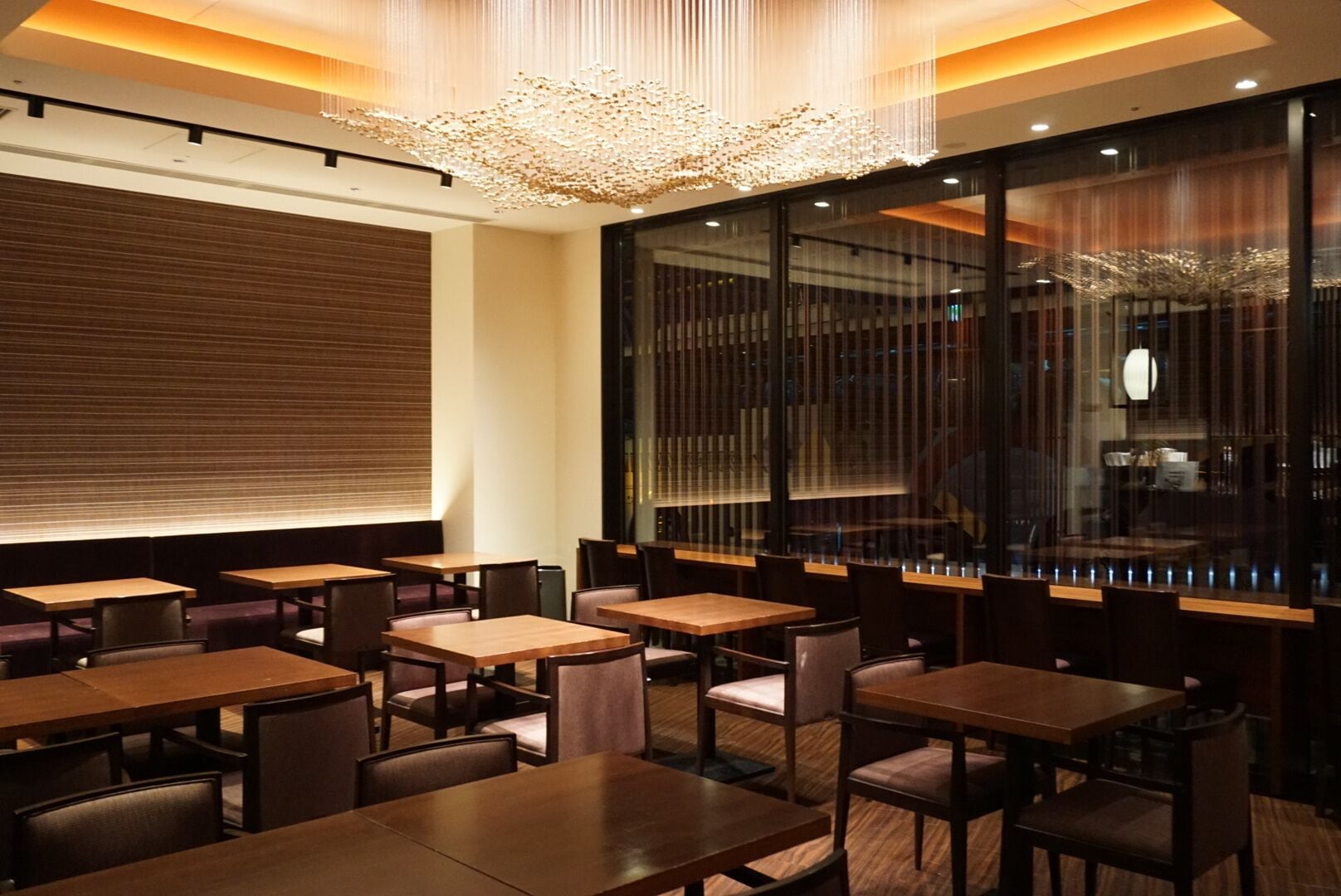 En modern restauranginteriör med träbord och stolar, en stor kristallkrona och utsikt genom ett fönster med ett dekorativt mönster.