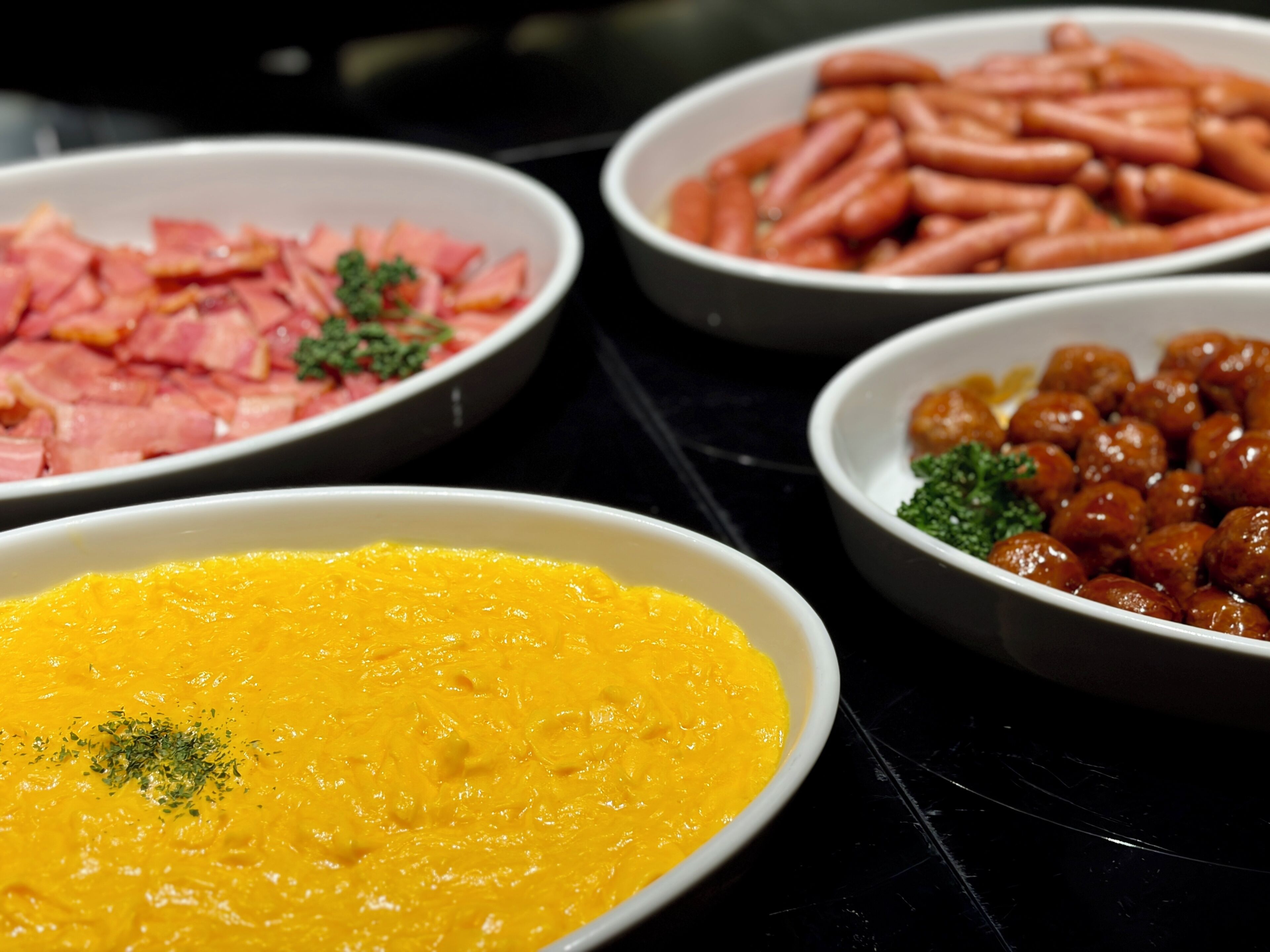 A buffet with various dishes including a bowl of yellow soup, a bowl of ham, a bowl of sausages, and a bowl of small round items garnished with parsley.