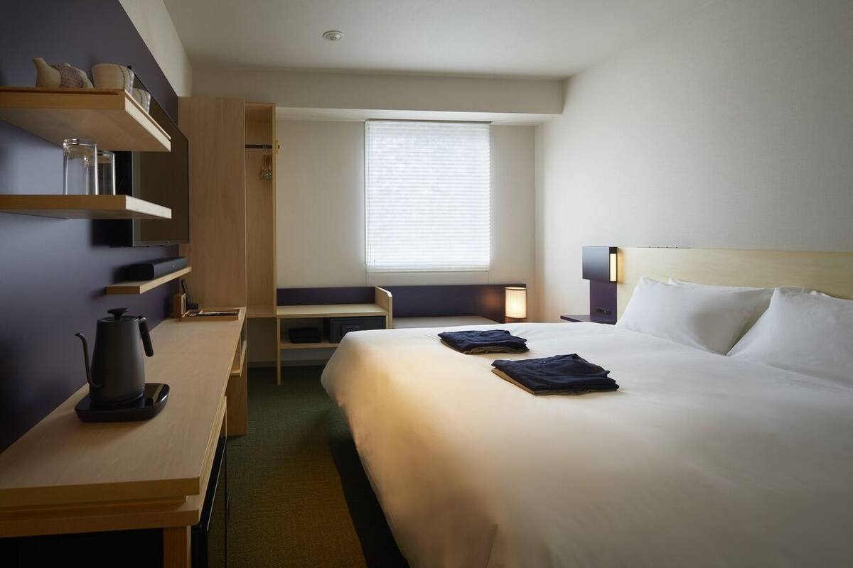 Ein Hotelzimmer mit Bett, Nachttisch, Schreibtisch, Stuhl, einem Fenster mit Jalousien und einem an der Wand befestigten Regal mit Gegenständen.