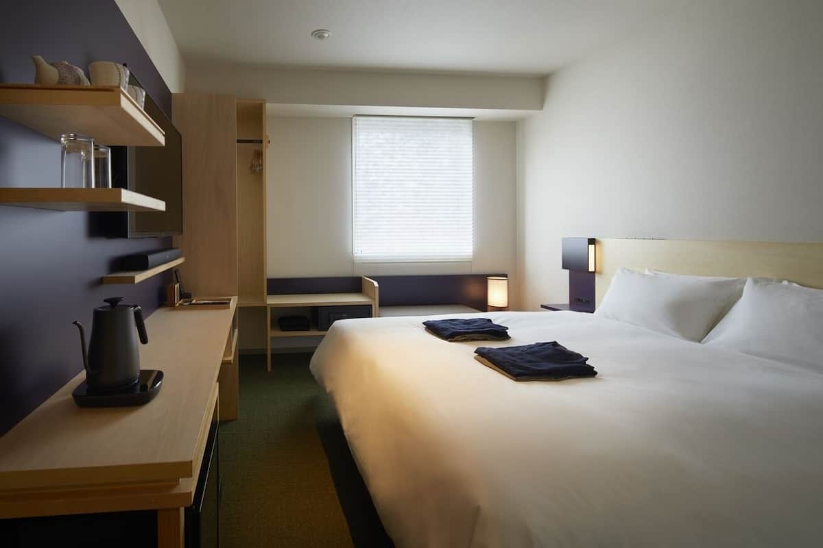Ein Hotelzimmer mit Bett, Nachttisch, Schreibtisch, Stuhl, einem Fenster mit Jalousien und einem an der Wand befestigten Regal mit Gegenständen.