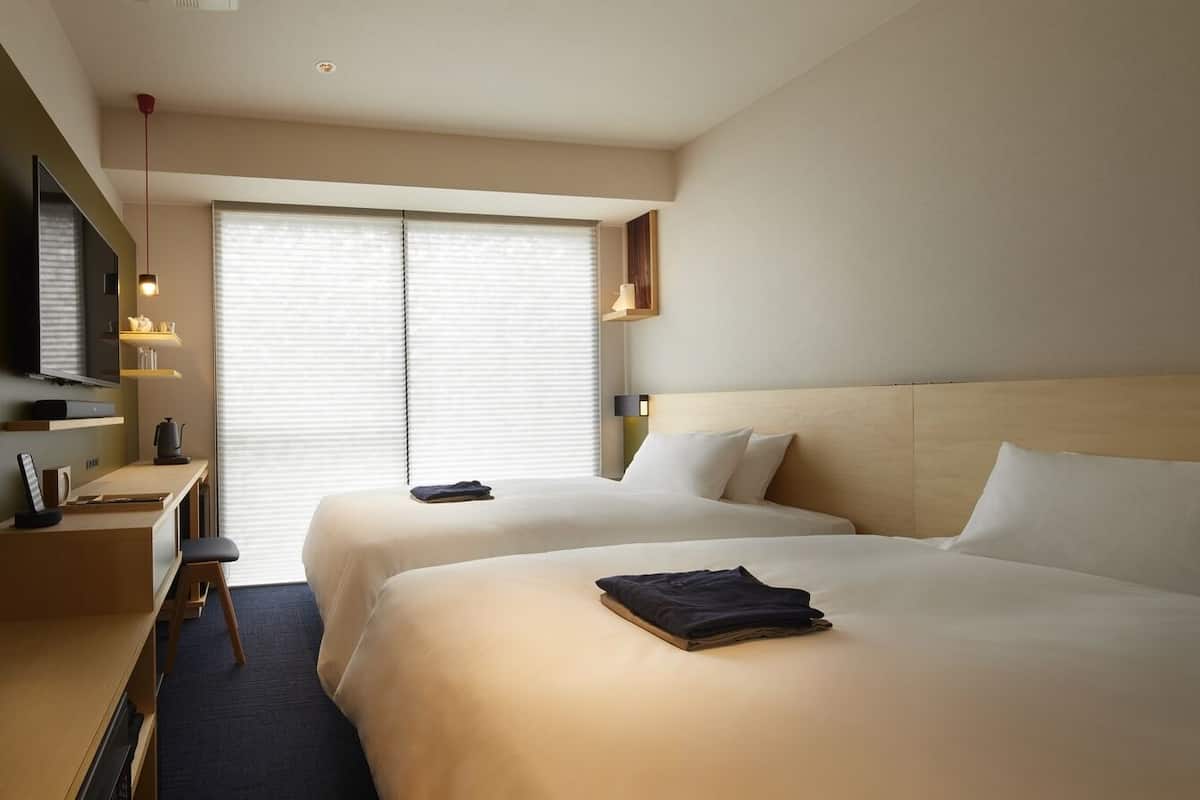 Ein Hotelzimmer mit zwei Betten, einem Schreibtisch, einem Fernseher und einem Fenster mit Jalousien.