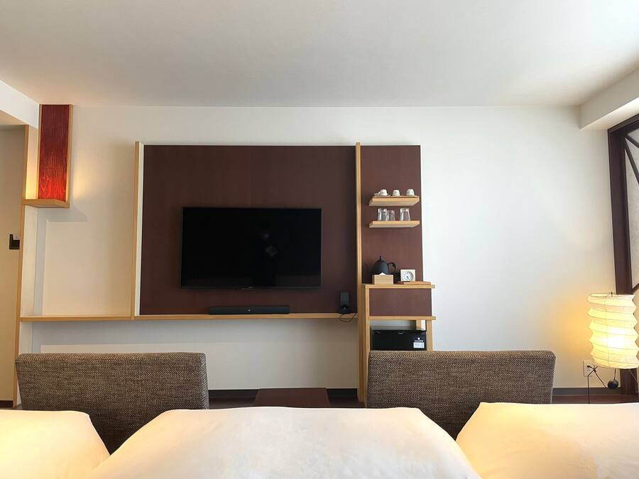 Et hotelværelse med et vægmonteret fladskærms-TV, en træhylde med pynteting, to polstrede lænestole og et natbord med en lampe.