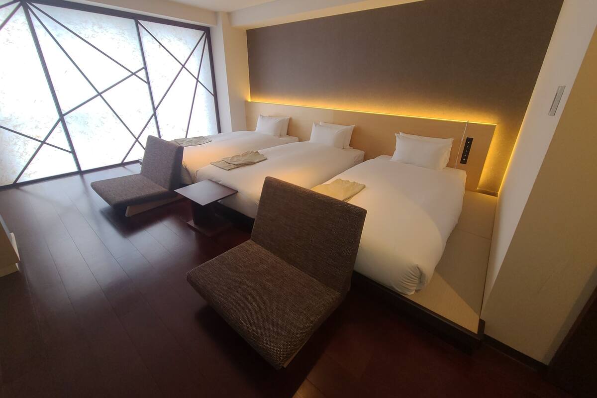 Ein Hotelzimmer mit zwei Betten, einem Schlafsofa, einem kleinen Tisch und einem großen Fenster mit geometrischem Muster.
