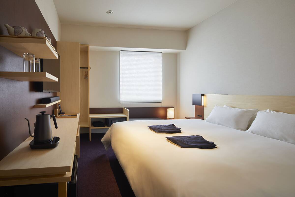Ein Hotelzimmer mit Bett, Schreibtisch mit Wasserkocher, Regalen mit Dekoration, Fenster mit Jalousien und Nachttisch mit Lampe.