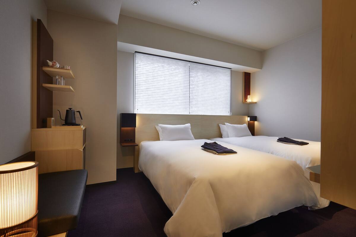 Ein Hotelzimmer mit zwei Betten, einem Nachttisch, einer Lampe und einem Fenster mit Jalousien.