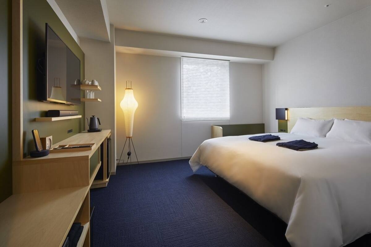 Ein Hotelzimmer mit Bett, Schreibtisch mit Lampe, Fernseher und Fenster mit Jalousien.