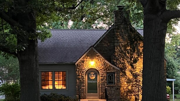 A stone cottage with a dark gray roof, a stone chimney, and a lit front door.
