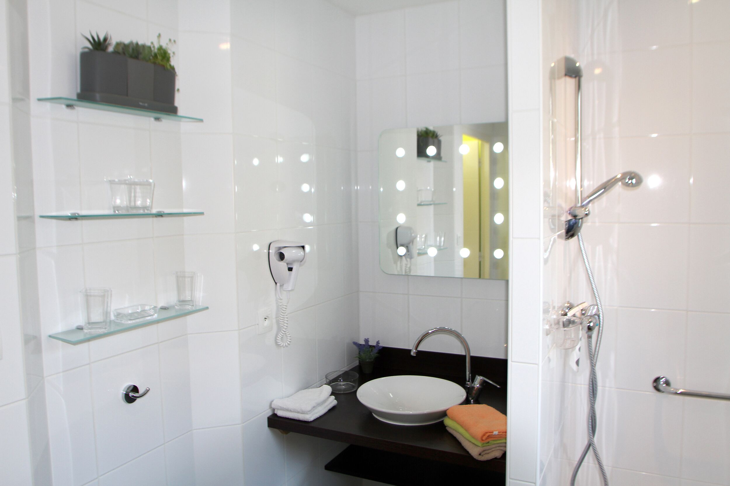 Ein modernes Badezimmer mit einer Glasablage, einem weißen Waschbecken, einem Spiegel mit Beleuchtung und einer Dusche.