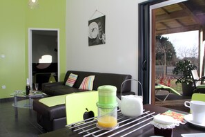A modern living room with a glass table, a sofa, and a dining area with a chair and a table set with a teapot, cups, and a fruit bowl.