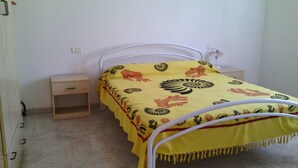 Un lit simple avec une couvre-lit jaune ornée de motifs floraux et animaliers, une tête de lit en métal blanche, une table de chevet avec un tiroir et une prise électrique au mur.