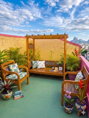 A wooden pergola with a cushioned bench, surrounded by potted plants and a view of a cityscape.