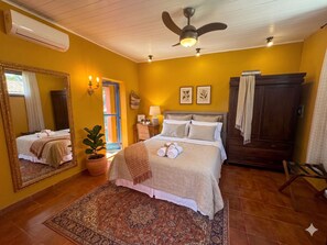 Ein Hotelzimmer mit einem Bett, einem Deckenventilator, einem Nachttisch, einer Lampe, einem Spiegel, einem Kleiderschrank und einer Zimmerpflanze.