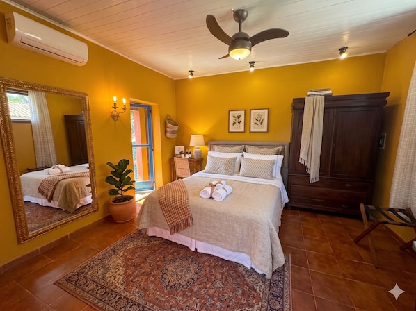 Ein Hotelzimmer mit einem Bett, einem Deckenventilator, einem Nachttisch, einer Lampe, einem Spiegel, einem Kleiderschrank und einer Zimmerpflanze.