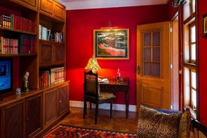 A room with a red wall, wooden bookshelves, a desk with a chair, a framed picture, and a door.