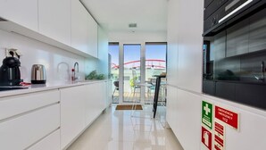 A modern kitchen with white cabinets, a coffee machine, a kettle, and a microwave. There are emergency signs on the wall and a view of a red bridge outside. - Lovage Gray Apartment, Benfica, Lisbon (Lisboa)