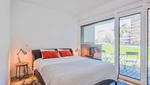 A bedroom with a large bed, a small side table, a lamp, and a view of the outside through a sliding glass door. - Lovage Gray Apartment, Benfica, Lisbon (Lisboa)