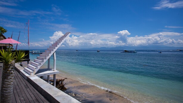 A wooden deck with a staircase leading to a beach, overlooking a calm sea with boats and clear skies. - Rijet Villa Beach & Restaurant (Penida Island)