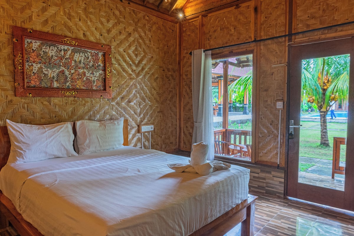 A room with a large bed, a wooden wall with a woven pattern, and a door leading to an outdoor area with a view of greenery.