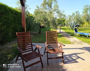 Two wooden chairs with slatted backs and armrests, placed on a tiled patio with a grassy area and trees in the background.