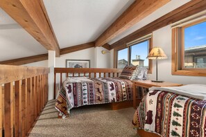 A bedroom with a wooden ceiling, a bed with a patterned blanket, a bedside table with a lamp, and a window with a view.