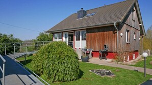 Ein zweistöckiges Haus mit rotem und weißem Anstrich, einer überdachten Veranda und einem kleinen Garten mit Buschwerk und Teich.