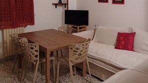 A living room with a wooden table and chairs, a television on a stand, a sofa with a red pillow, and two framed pictures on the wall.