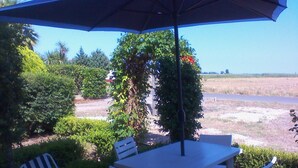 An outdoor patio with a table and chairs under a large umbrella, surrounded by lush greenery and a view of a water body.