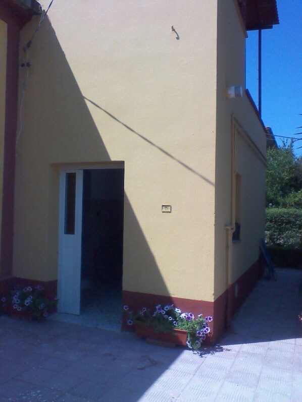Un edificio con fachada amarilla, una puerta y un pequeño ventanal. Hay macetas con flores púrpuras a la base del edificio.