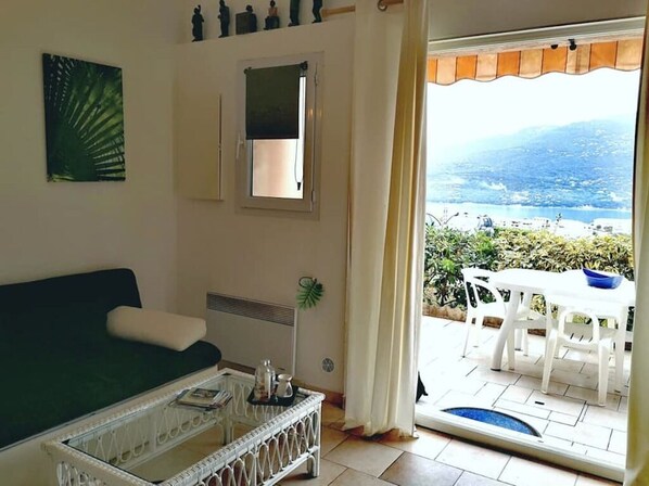 A living room with a sofa, a coffee table, and a view of an outdoor patio with a table and chairs. - Large studio with stunning sea view for breakfast and 5 minutes from the beach (Propriano)