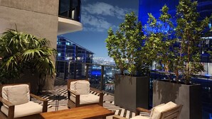 Una terraza en la azotea con mobiliario exterior, plantas en macetas y vistas a la ciudad de noche.