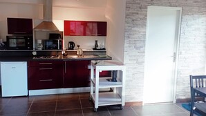 Una cocina moderna con armarios rojo oscuro, una encimera blanca y un horno empotrado. Hay un carro pequeño blanco con un hervidor negro y una cafetera. La cocina tiene piso de baldosas y una puerta blanca a la derecha.