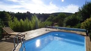 Una terraza de madera con piscina, rodeada de abundante vegetación y colinas al fondo.