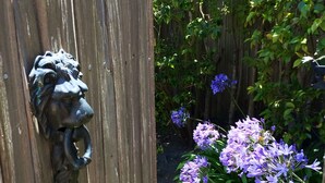 Una verja de madera con un tirador en forma de cabeza de león, rodeada de abundante follaje y flores moradas.