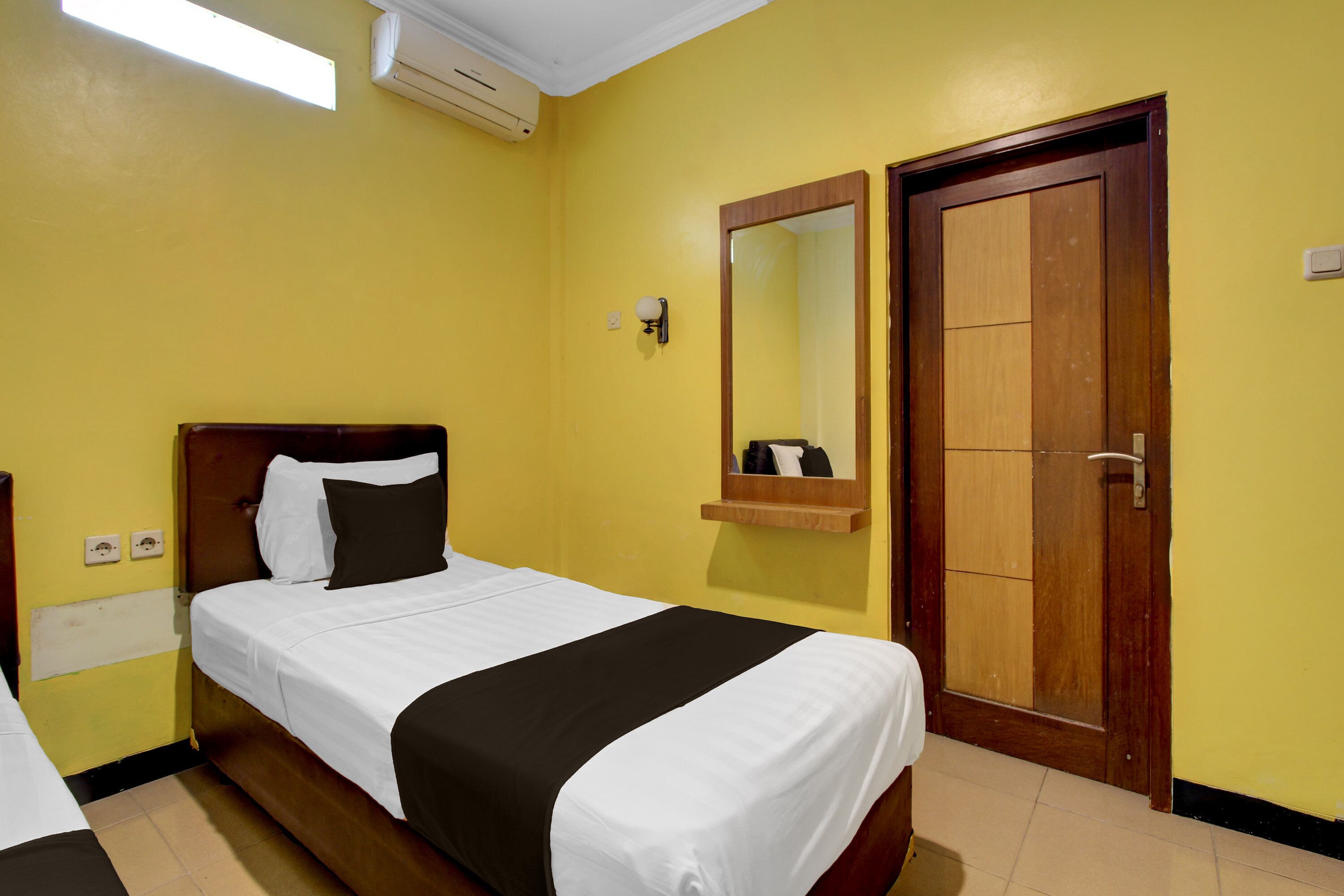 A hotel room with a bed, a wooden door, a mirror, and an air conditioning unit.