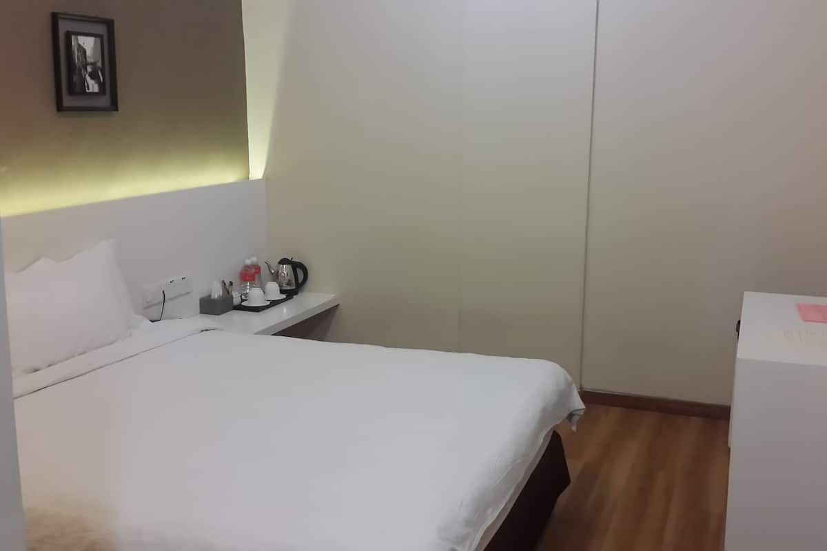 A hotel room with a bed, a small table with a kettle, cups, and a water bottle. There is a TV on the wall and a whiteboard with a note.