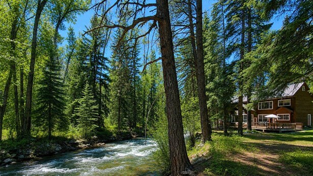 Una baita sul fiume con un patio, circondata da una fitta foresta e un ruscello cristallino.