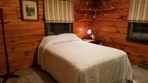 A wooden cabin bedroom with a bed, a nightstand, a lamp, and a shelf with figurines. - Log cabin nestled beside Chiques Creek in historic Lancaster County (Mount Joy)