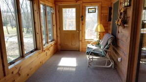 A cozy cabin interior with a wooden door, large windows, a chair, a lamp, and a small table. - Log cabin nestled beside Chiques Creek in historic Lancaster County (Mount Joy)