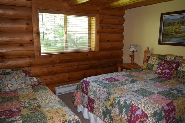 A log cabin bedroom with two beds, a window with blinds, a nightstand with a lamp, and a painting on the wall.