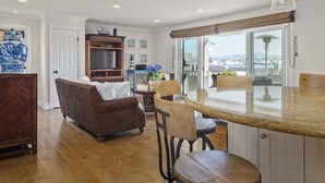 A kitchen with a large island, a dining area with chairs, a wicker sofa, and a view through a window. - LUX Beachfront Casa de Balboa 231 w/AC - Best View (Newport Beach)
