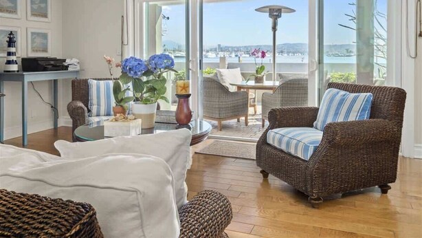 A living room with a wicker sofa, a white couch, and a dining table with chairs. There is a large window with a view of the sea and boats. - LUX Beachfront Casa de Balboa 231 w/AC - Best View (Newport Beach)