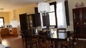 A dining area with a dark wooden table, six chairs, and a chandelier. There is a kitchen in the background with wooden cabinets and a window with curtains. - Rural villa in Marrucho Alto (Riogordo)