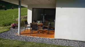 Un patio exterior moderno con una mesa y sillas, rodeado por un borde de grava y una zona verde con vacas al fondo.