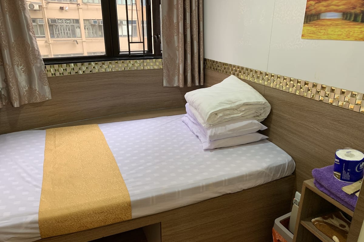 A single bed with a wooden headboard, a patterned bedspread, and a neatly folded white pillow. A small trash bin is visible to the right of the bed.